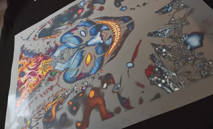 Feuille de transfert DTF vue en biais sur un vêtement foncé : un masque bleu, des flammes et plusieurs petits motifs imprimés côte à côte sur le même film.